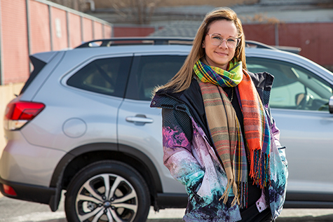 Image of woman with glasses in a multicolored scarf standing in front of a silver vehicle