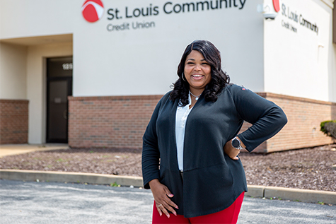 Image of lady in white shirt with red pants standing in front of Slccu building smiling and posing with hand on hip.